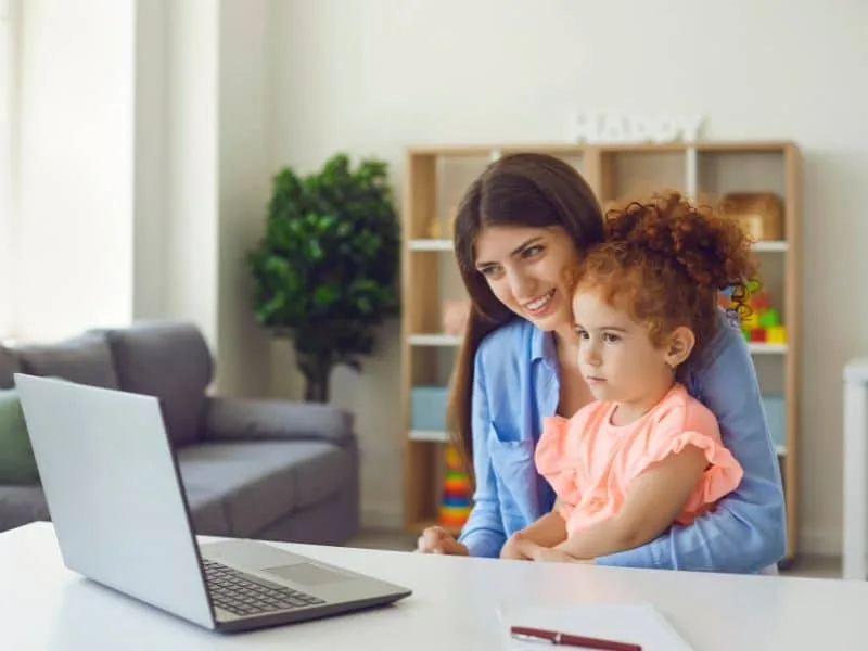 A young child on a video call with friends while a nanny supervises.